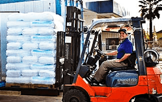 A person operates an orange forklift, lifting a pallet stacked with large bags for an ice delivery in front of a blue building.