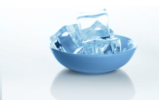 A small blue bowl filled with clear ice cubes is placed on a reflective white surface.