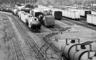 Black and white image of a train yard with multiple tracks and various freight cars, including tankers and boxcars arranged in parallel lines, reminiscent of goods delivered before refrigerators became commonplace.