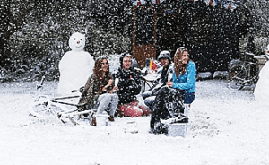 Four people are sitting in the snow, holding beverages, with a snowman and bicycles in the background on a snowy day.