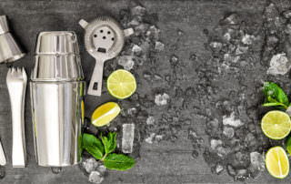 A cocktail shaker, strainer, jigger, stirring spoon, muddler, and lime slices are arranged on a dark surface with ice cubes and mint leaves, ready to craft the perfect cocktails.