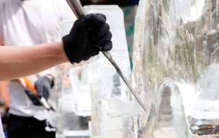 A sculptor wearing black gloves meticulously carves an ice sculpture using chisels. Another person in the background also works on the intricate creation.
