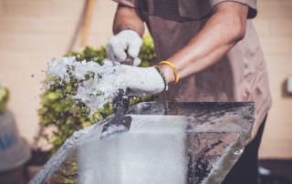 A person wearing gloves meticulously carves a block of ice with a chisel, causing ice fragments to scatter.