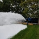 A person operates a foam machine, covering an area of grass and hay bales with foam, while an ice delivery truck is parked nearby amidst the trees in the background.