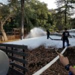 Two people spray artificial snow onto a wooded area while another person looks at their phone in the foreground. Nearby, equipment and a large hose for ice delivery are visible.