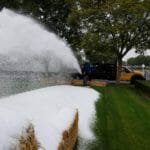 A person operates a machine spraying artificial snow over a row of hay bales on a grassy area, with trees in the background, creating an atmosphere reminiscent of an ice delivery service.