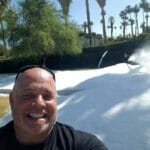 A man smiles at the camera while another person surfs on artificial snow in a sunny area with palm trees, hinting at the refreshing touch brought by recent ice delivery.