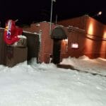 A snow-covered alley beside a brick building with lights on. A red cloth with colorful text hangs over a brown fence next to the building, hinting at an ice delivery service. Snow has accumulated on the ground and on the fence.