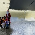 A group of children poses with a person dressed as Santa Claus under a canopy next to a large pile of artificial snow, reminiscent of an ice delivery gone festive.