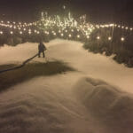 A person operates a snow machine, covering a yard with artificial snow at night. Amid the scene, string lights hang above, casting a warm glow while an ice delivery awaits nearby.