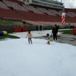 Two children play on artificial snow on a stadium field, where ice delivery had been arranged for a special event. An adult stands nearby, overseeing the fun. The stands in the background are mostly empty, with red and white balloons floating in the distance.