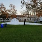 A park scene shows a grassy area where people stand near a mound of artificial snow, with trash bins, a white tent, and a Ryder truck in the background. An ice delivery has just taken place, adding to the festive atmosphere. Trees and buildings surround the scene.