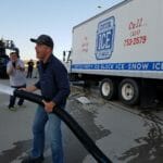 A person in a blue shirt and cap holding a large hose, standing near a truck labeled "Crystal Ice," appears to be managing an ice delivery. Other individuals and equipment are visible in the background.