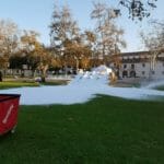 A grassy area is covered with a large amount of foam and surrounded by trees and buildings. A red bin is on the left, and a white tent for ice delivery stands near a fountain in the background.